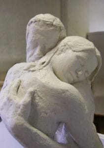 Close-up detail of 'Peaceful Embrace', a bonded marble sculpture. One figure rests their head on the other's shoulder, eyes closed. The textured surface of the work is visible.