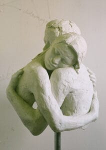 'Peaceful Embrace' bonded marble sculpture, front view. Two figures embrace, both with closed eyes and serene expressions, their forms merging at the center. Mounted on a metal rod.