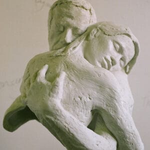 White bonded marble sculpture of two figures embracing. Both have their eyes closed and serene expressions. Their bodies are cut off at mid-chest, and where they meet, their forms dissolve into one another.