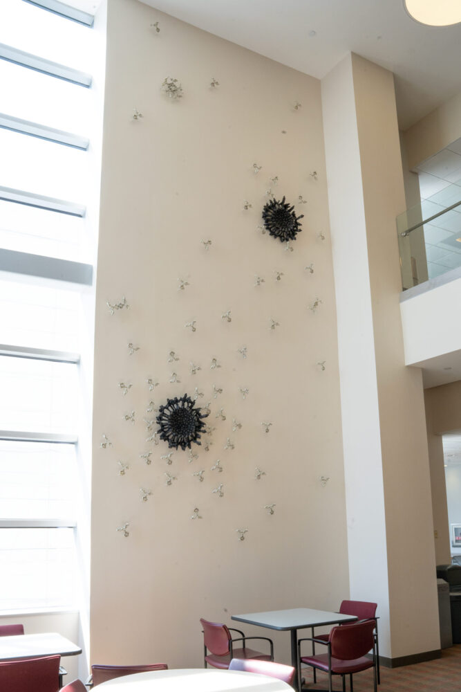 Full view of Silent Sentinels antibody sculpture by Briony Marshall, photographed from the left side. The relief sculpture installation covers a large wall measuring 3m wide by 7.5m tall (10' x 24'), featuring two dark ominous viruses being swarmed by silver Y-shaped antibodies cast in bonded bronze and pewter, in a classic battle of good versus evil. A large window is visible on the left. A balcony is visible on the right, with seating and tables visible below the installation. Installed outside the Crowe Lab at Vanderbilt University Medical Centre in Nashville.